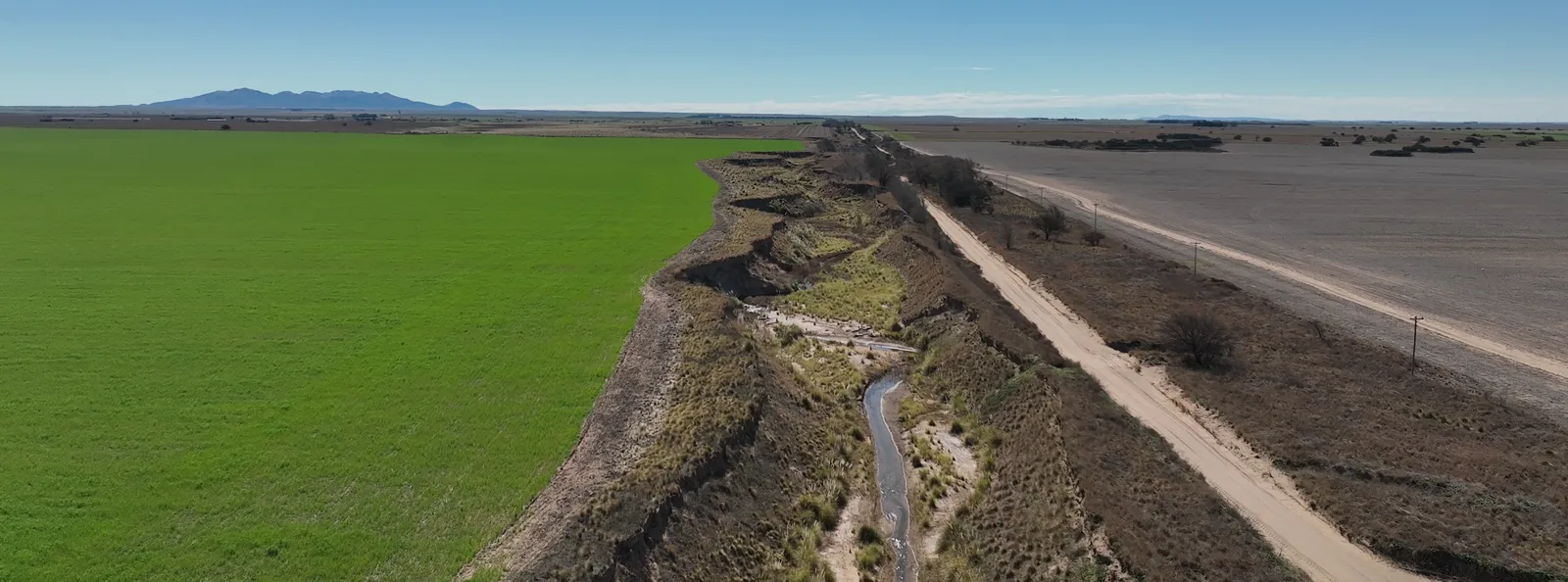 Vista aérea oblicua desde el dron: un lote verde, una cárcava con agua en el medio y un camino de tierra, con las sierras al fondo